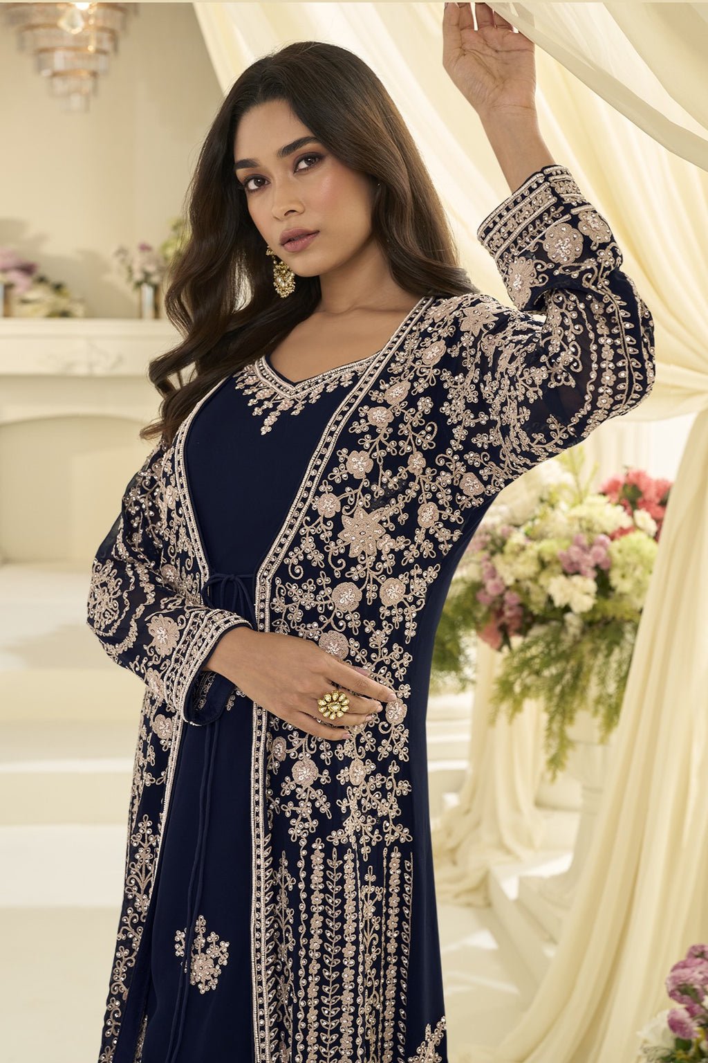 A woman models a navy blue traditional outfit with extensive  embroidery, set against a warm, cream-colored background with floral arrangements.