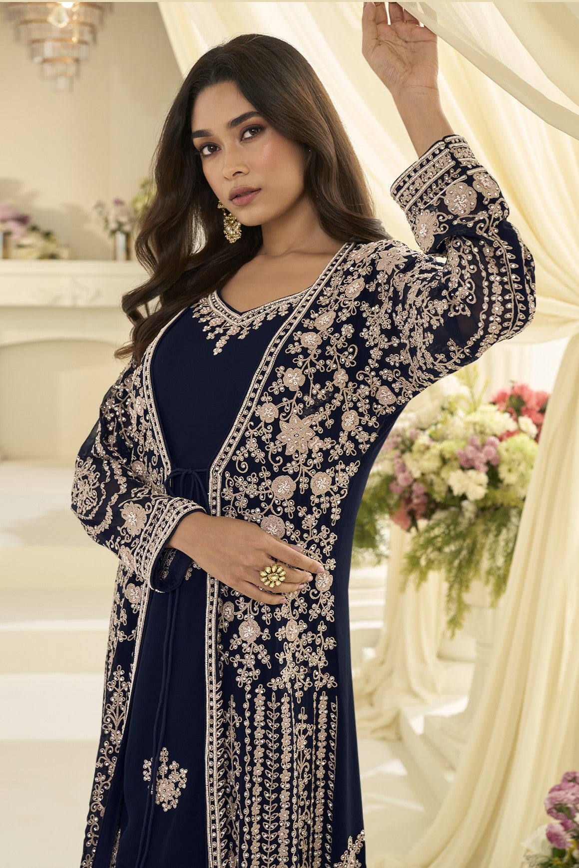 A woman models a navy blue traditional outfit with extensive  embroidery, set against a warm, cream-colored background with floral arrangements.