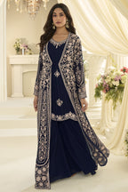 A woman models a traditional South Asian outfit featuring a navy blue dress with intricate embroidery, paired with a matching long open-front jacket, set against an elegant cream-colored indoor backdrop.
