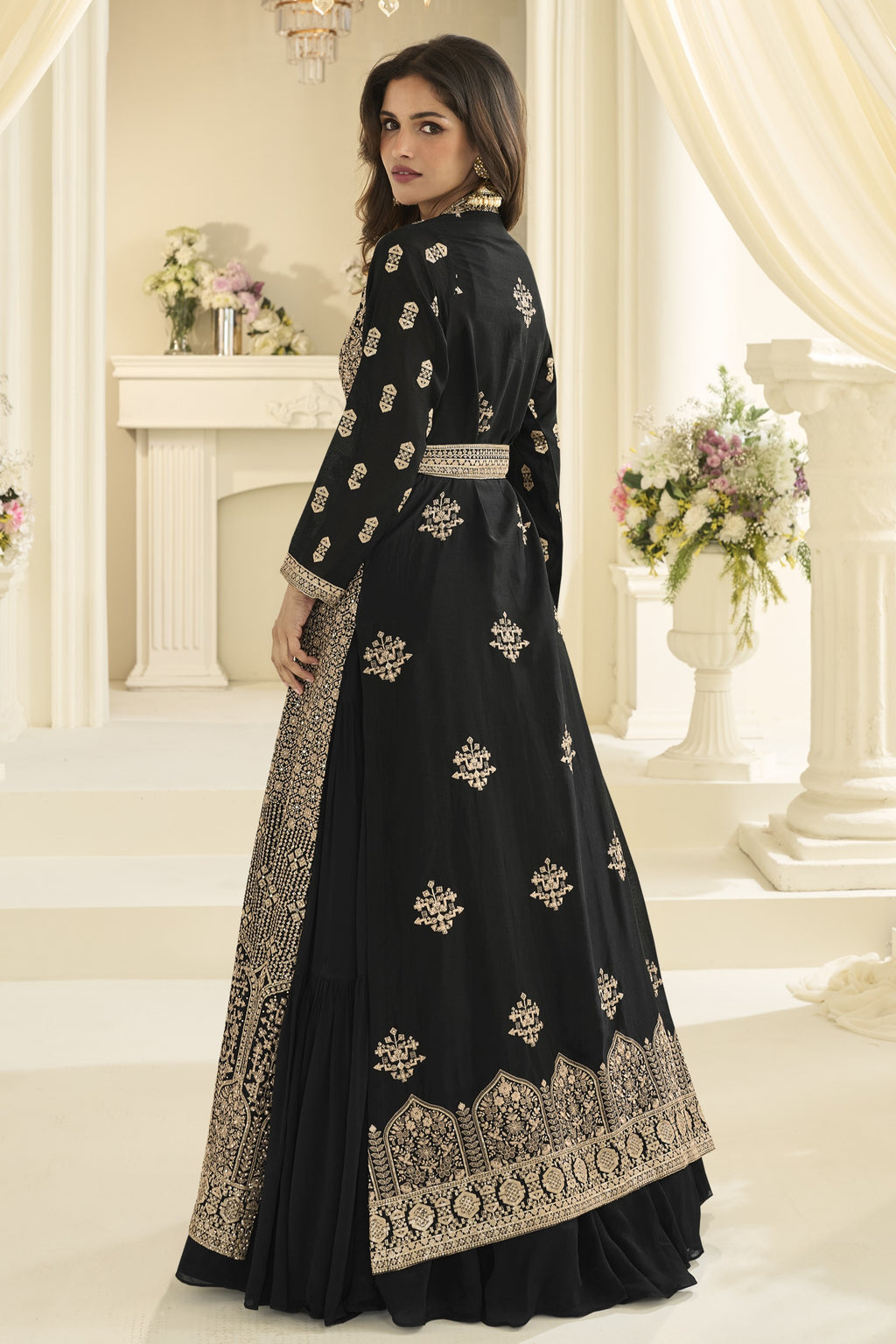 A woman models a black traditional long gown with extensive gold and silver embroidery, set against an elegant cream-colored classical interior backdrop.