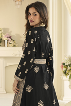 A woman looking back and wearing a black traditional outfit with intricate embroidery and a matching belt, photographed in an elegant indoor setting with a cream-colored fireplace and floral arrangements.