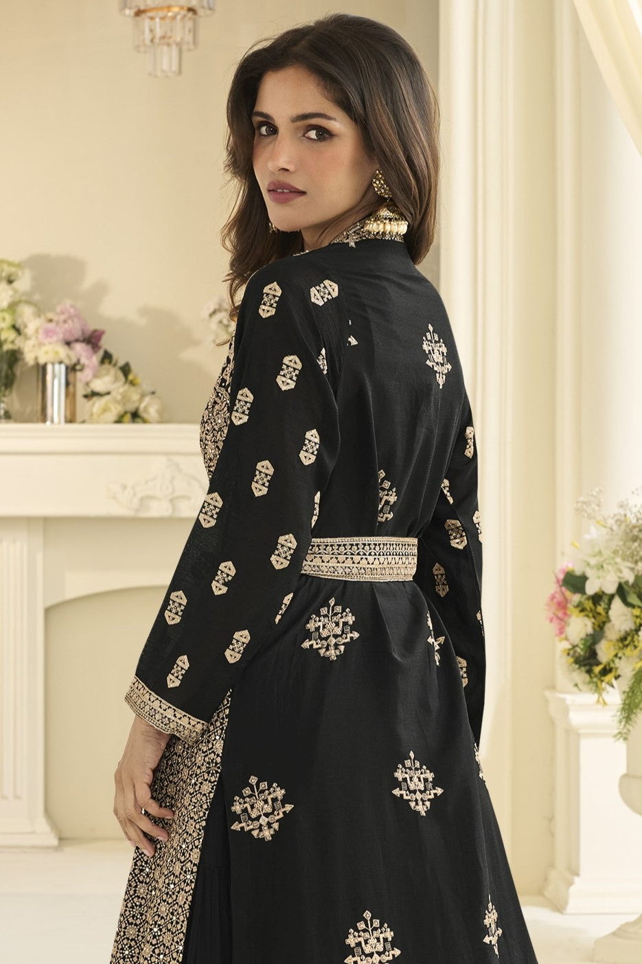 A woman looking back and wearing a black traditional outfit with intricate embroidery and a matching belt, photographed in an elegant indoor setting with a cream-colored fireplace and floral arrangements.