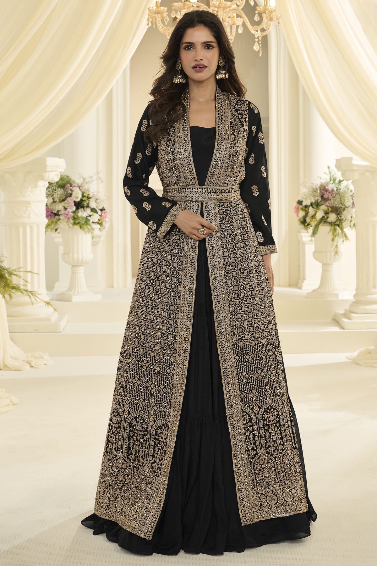 A woman models a long black dress with extensive embroidery, styled with a matching embroidered open-front jacket. The setting is an elegant indoor space with draped cream curtains, columns, and floral arrangements.