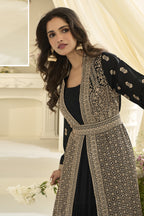 A woman models a black fabric jacket with embroidery and a matching embroidered belt, set against a warm cream-colored indoor backdrop.
