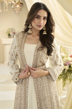 A woman models a traditional South Asian outfit featuring a cream-colored anarkali with intricate embroidery, worn under a matching open-front embroidered jacket. She is accessorized with large gold earrings and a gold ring, set against a warm, elegant indoor backdrop with draped cream fabric.