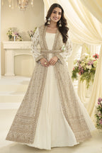 A woman models a traditional South Asian outfit featuring a cream floor-length anarkali gown with a long, heavily embroidered outer coat/jacket. The setting is a beautifully decorated indoor space with draped cream fabric, a white fireplace mantel, and floral arrangements.