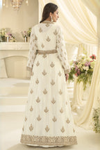 A woman wearing a long cream-colored traditional anarkali with embroidery, shown from the back in an elegant indoor setting with draped cream fabric and floral arrangements.