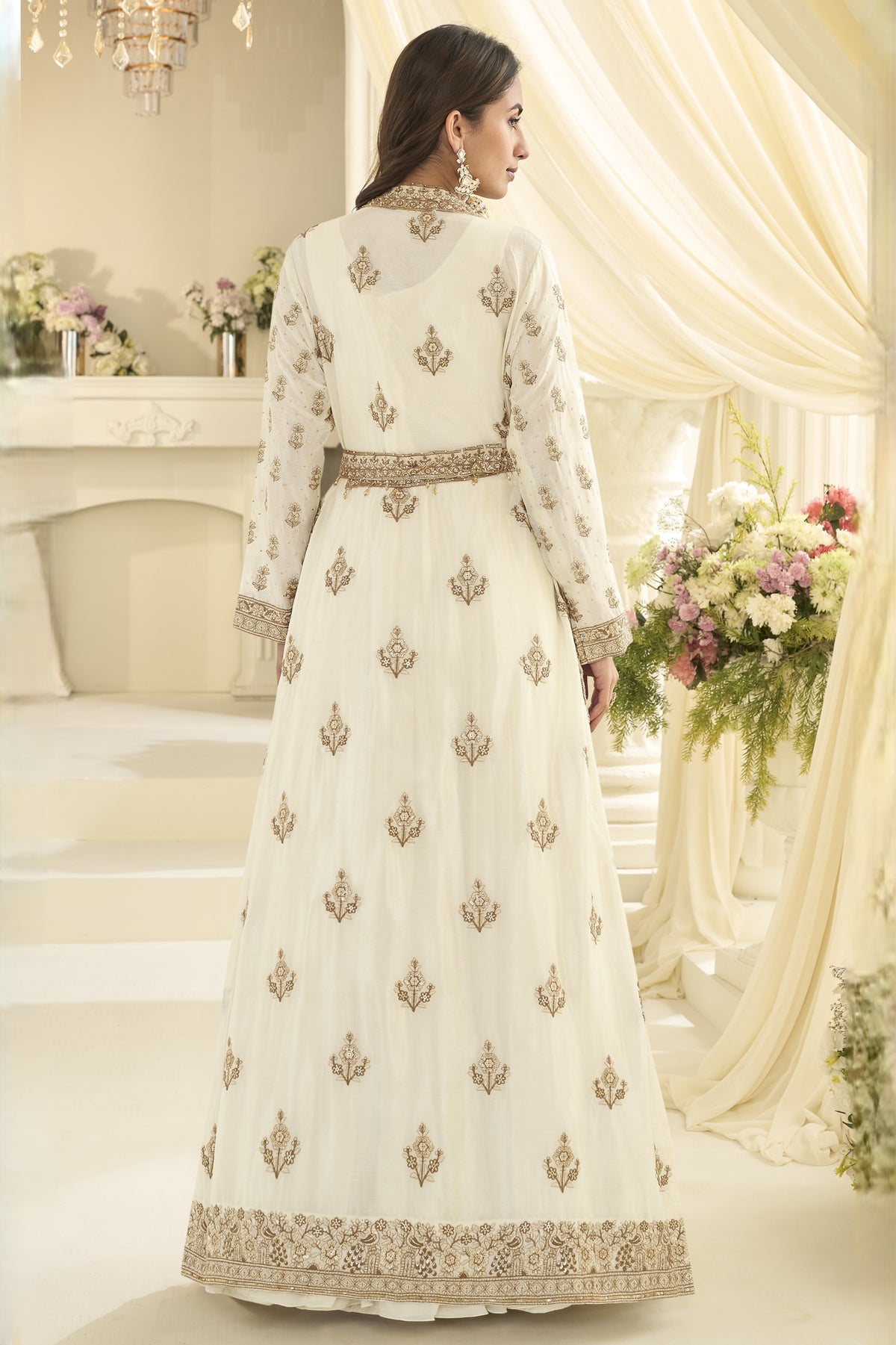 A woman wearing a long cream-colored traditional anarkali with embroidery, shown from the back in an elegant indoor setting with draped cream fabric and floral arrangements.