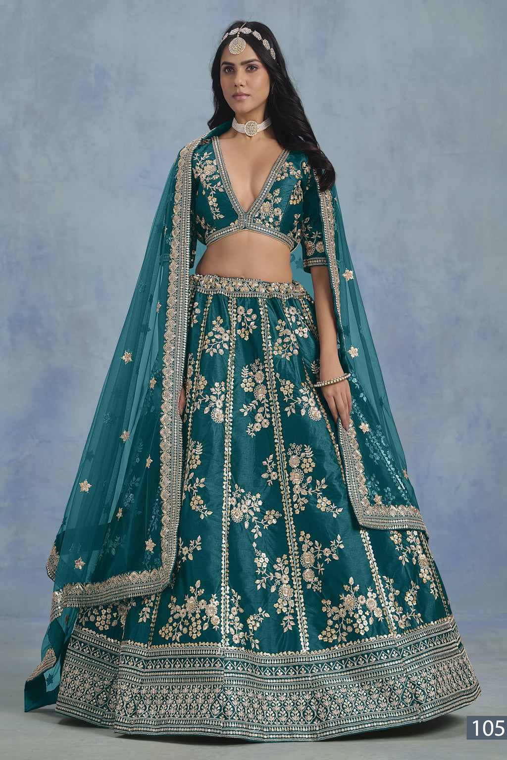 A woman in a full-length front view wears a Teal blue embroidered lehenga choli for reception with floral work and net dupatta on a blue studio backdrop.