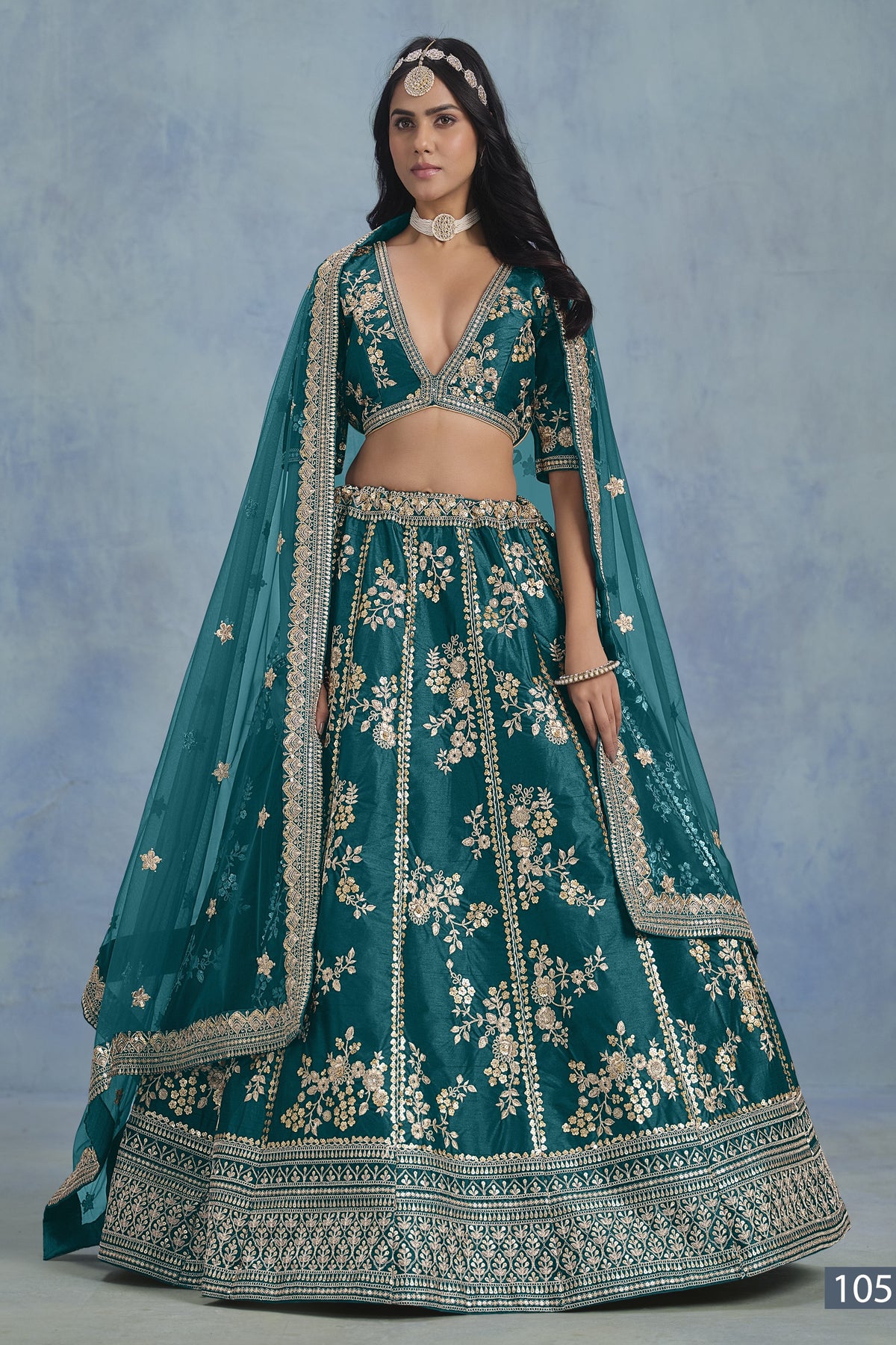 A woman in a full-length front view wears a Teal blue embroidered lehenga choli for reception with floral work and net dupatta on a blue studio backdrop.