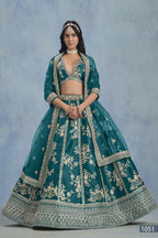 A woman faces forward in a full-length Teal blue embroidered lehenga choli for reception, with embroidered skirt and net dupatta on a blue backdrop.