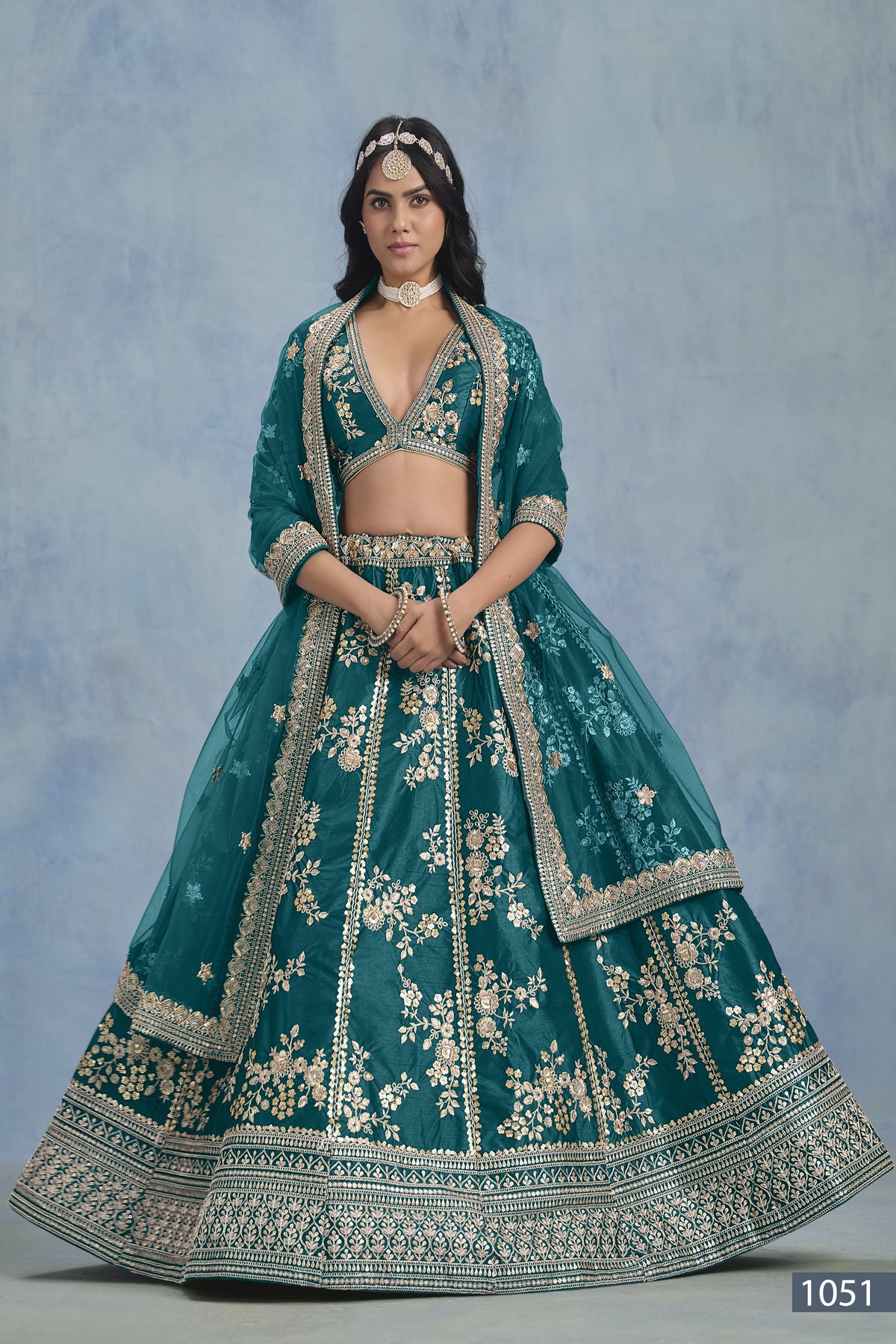 A woman faces forward in a full-length Teal blue embroidered lehenga choli for reception, with embroidered skirt and net dupatta on a blue backdrop.