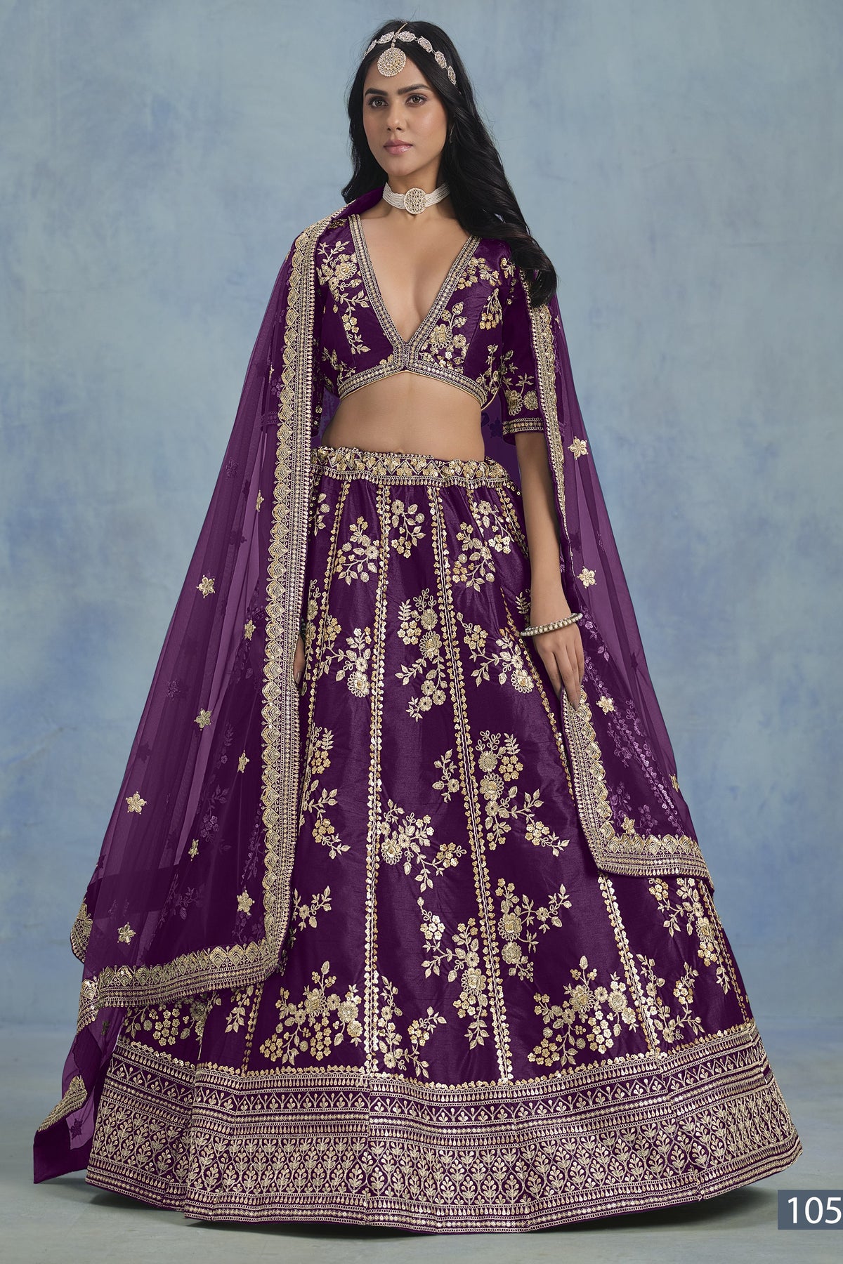 A woman in a full-length front view wears a purple embroidered lehenga choli for reception with floral work and net dupatta on a blue studio backdrop.