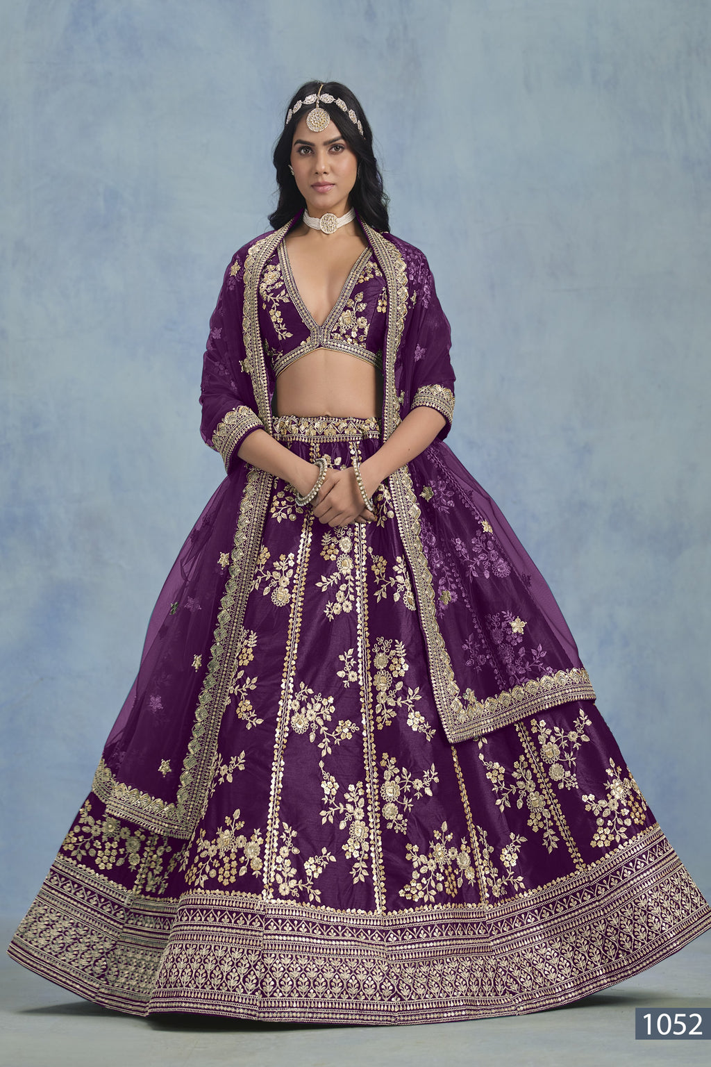 A woman faces forward in a full-length purple embroidered lehenga choli for reception, with embroidered skirt and net dupatta on a blue backdrop.