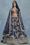 A woman in a full-length front view wears a navy blue embroidered lehenga choli for reception with floral work and net dupatta on a blue studio backdrop.