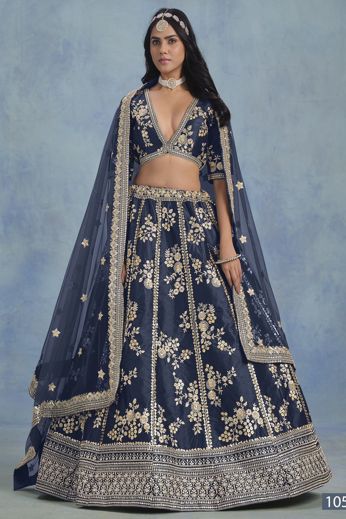 A woman in a full-length front view wears a navy blue embroidered lehenga choli for reception with floral work and net dupatta on a blue studio backdrop.