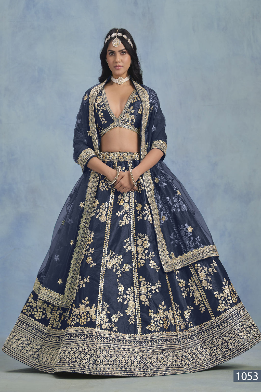 A woman faces forward in a full-length navy blue embroidered lehenga choli for reception, with embroidered skirt and net dupatta on a blue backdrop.
