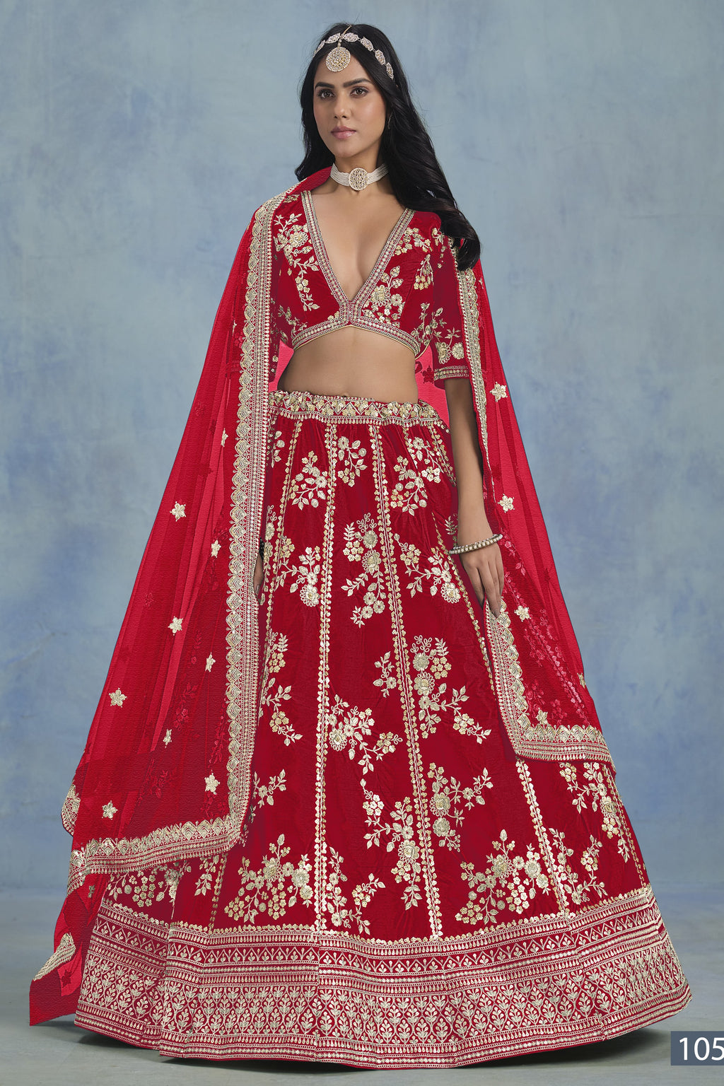 A woman in a full-length front view wears a red embroidered lehenga choli for party with floral work and net dupatta on a blue studio backdrop.