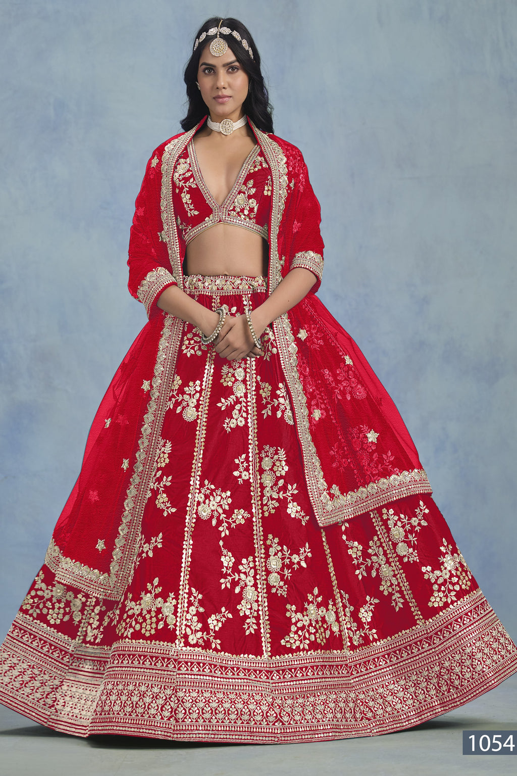 A woman faces forward in a full-length red embroidered lehenga choli for party, with embroidered skirt and net dupatta on a blue backdrop.