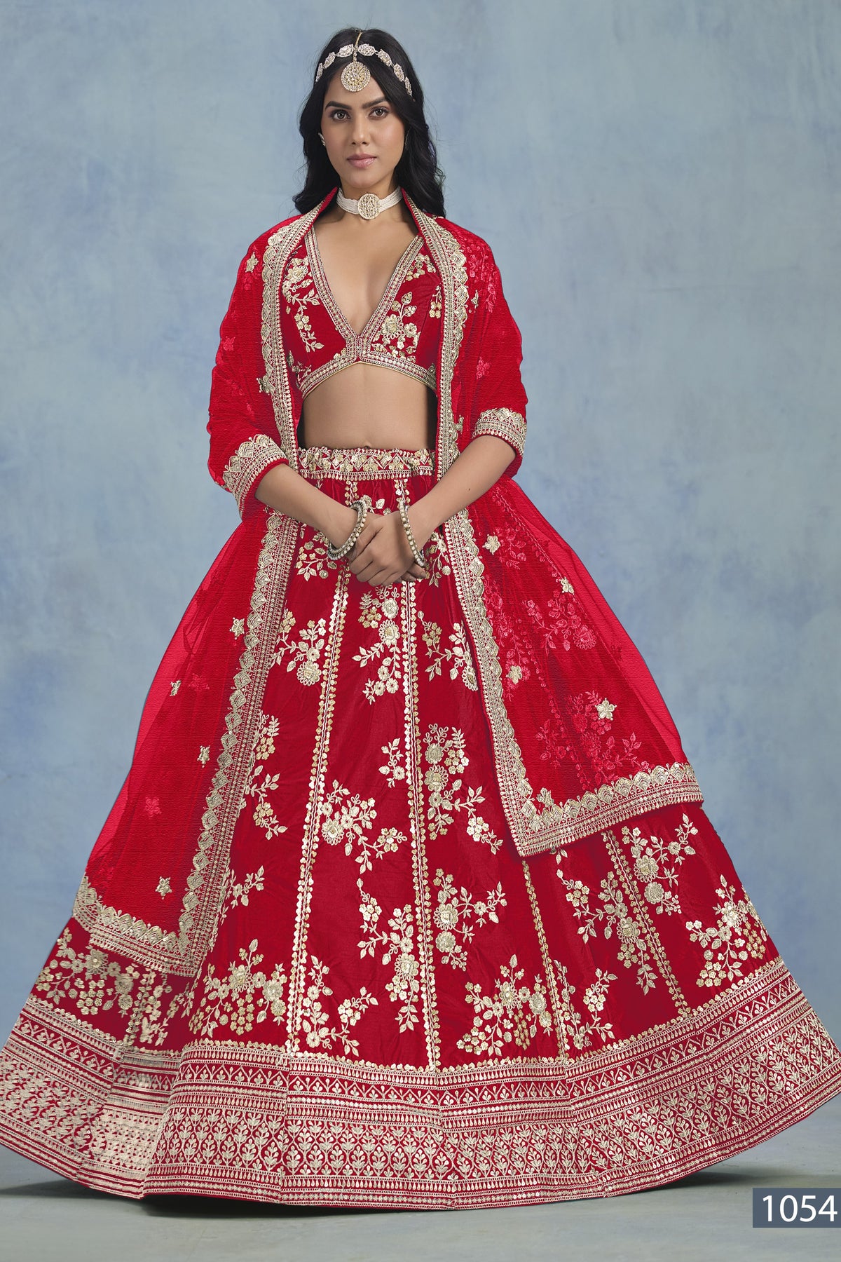 A woman faces forward in a full-length red embroidered lehenga choli for party, with embroidered skirt and net dupatta on a blue backdrop.