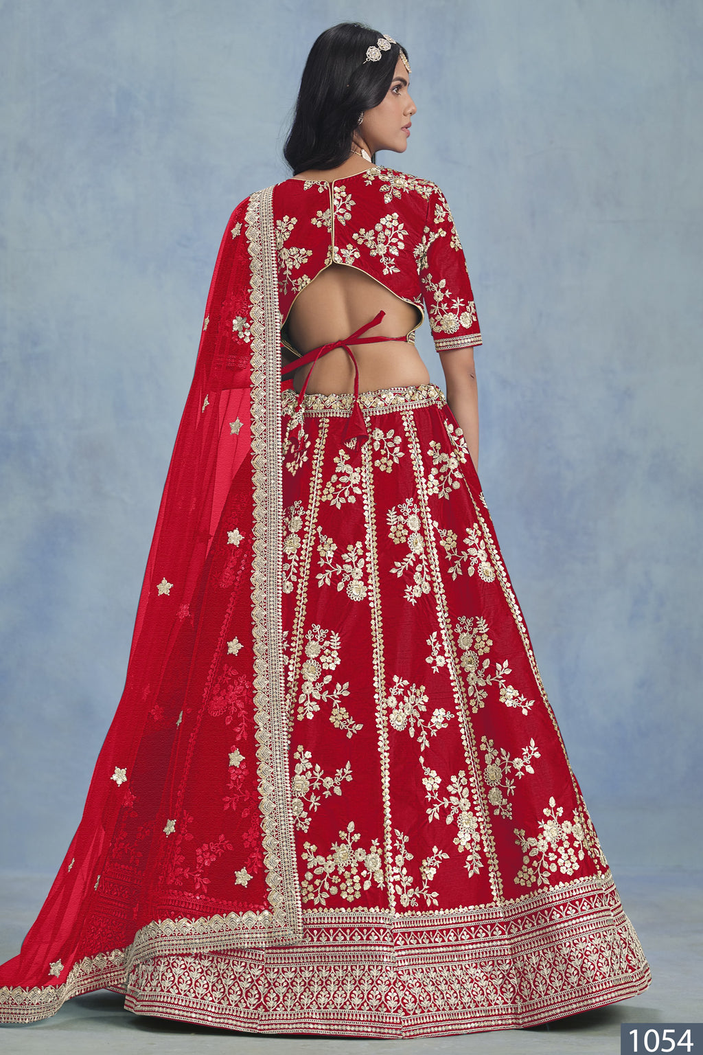 A back view of a woman in a red embroidered lehenga choli for party shows the open-back blouse, embroidered skirt and net dupatta.