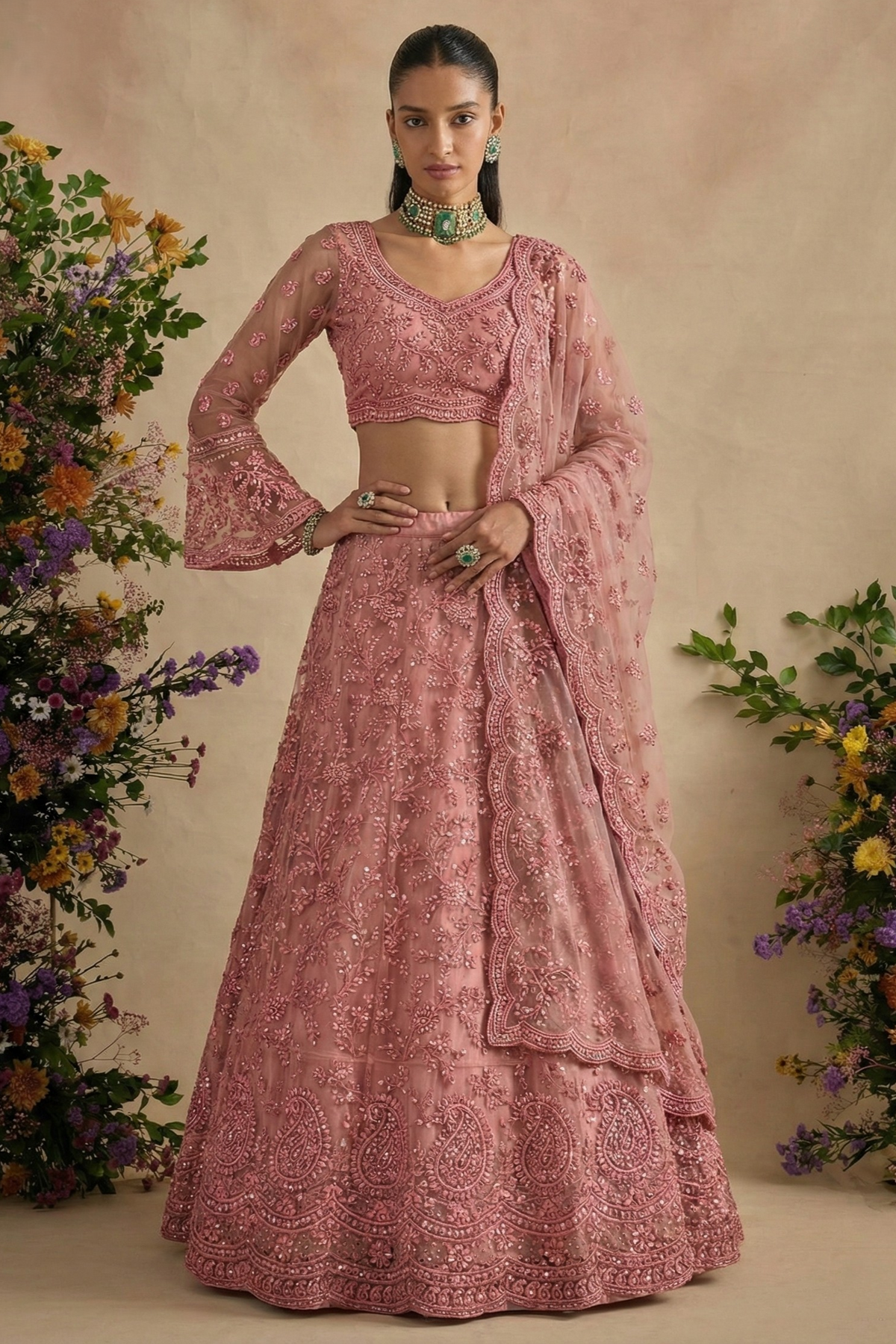Model wearing a dusty pink net lehenga choli with bell sleeves, V neckline and scalloped dupatta, hand on hip — a charming engagement lehenga with stone work.