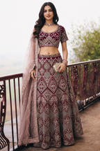 A woman stands on an outdoor balcony wearing an embroidered Indian ethnic outfit consisting of a heavily embellished maroon  choli, a matching flared skirt with geometric patterns, and a sheer dupatta.
