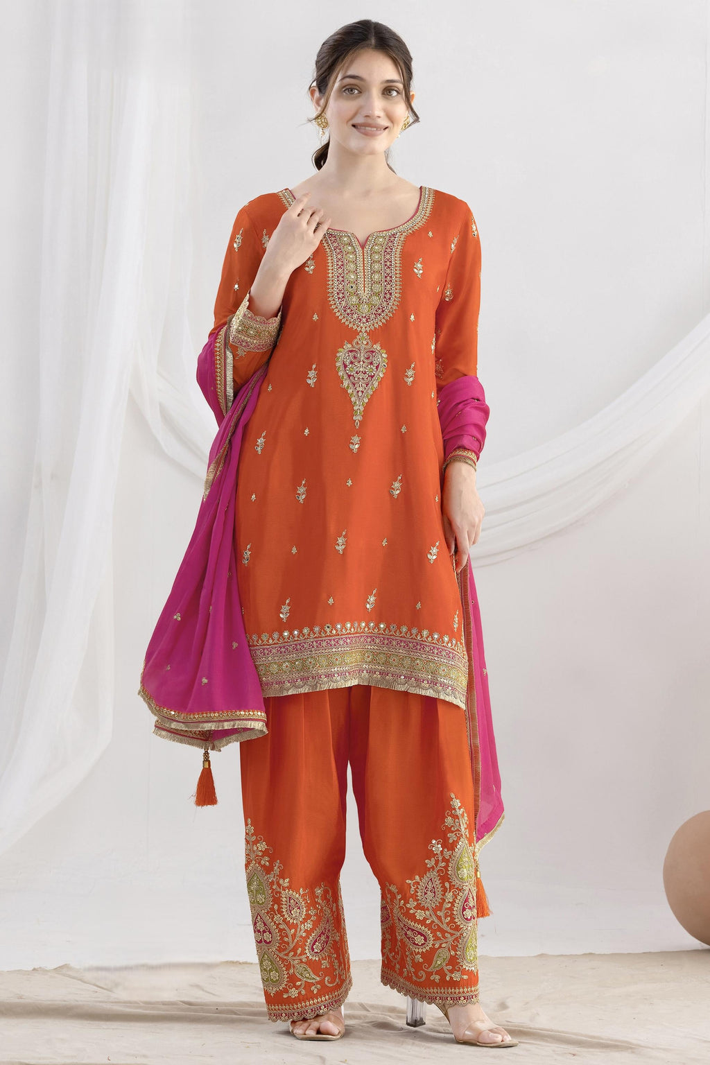 Woman wearing an orange embroidered traditional outfit with a pink dupatta on a white background