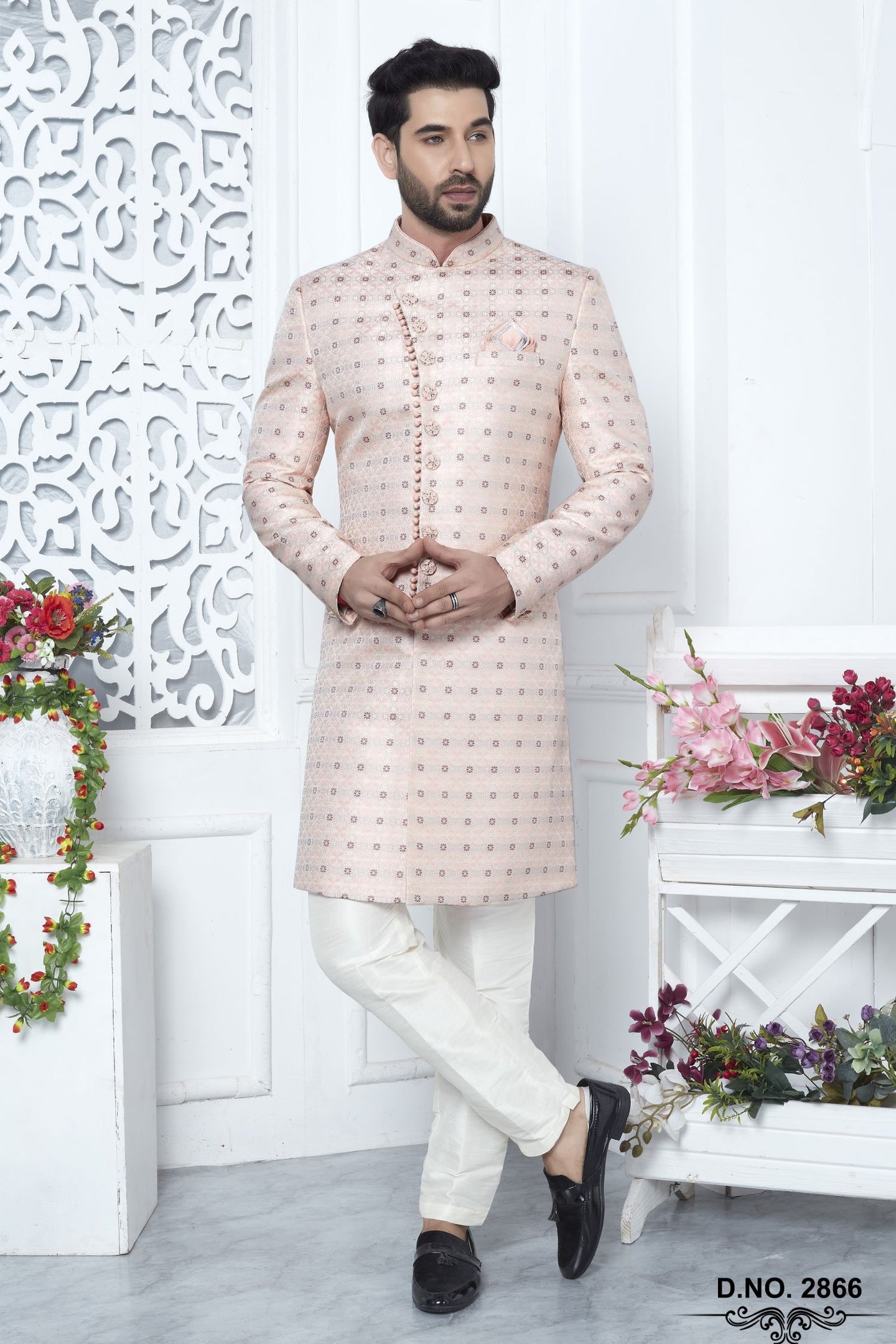 A man modeling a light beige sherwani with a subtle pink and green floral pattern, paired with white trousers and black shoes, standing in a decorated indoor setting with white walls and floral arrangements.