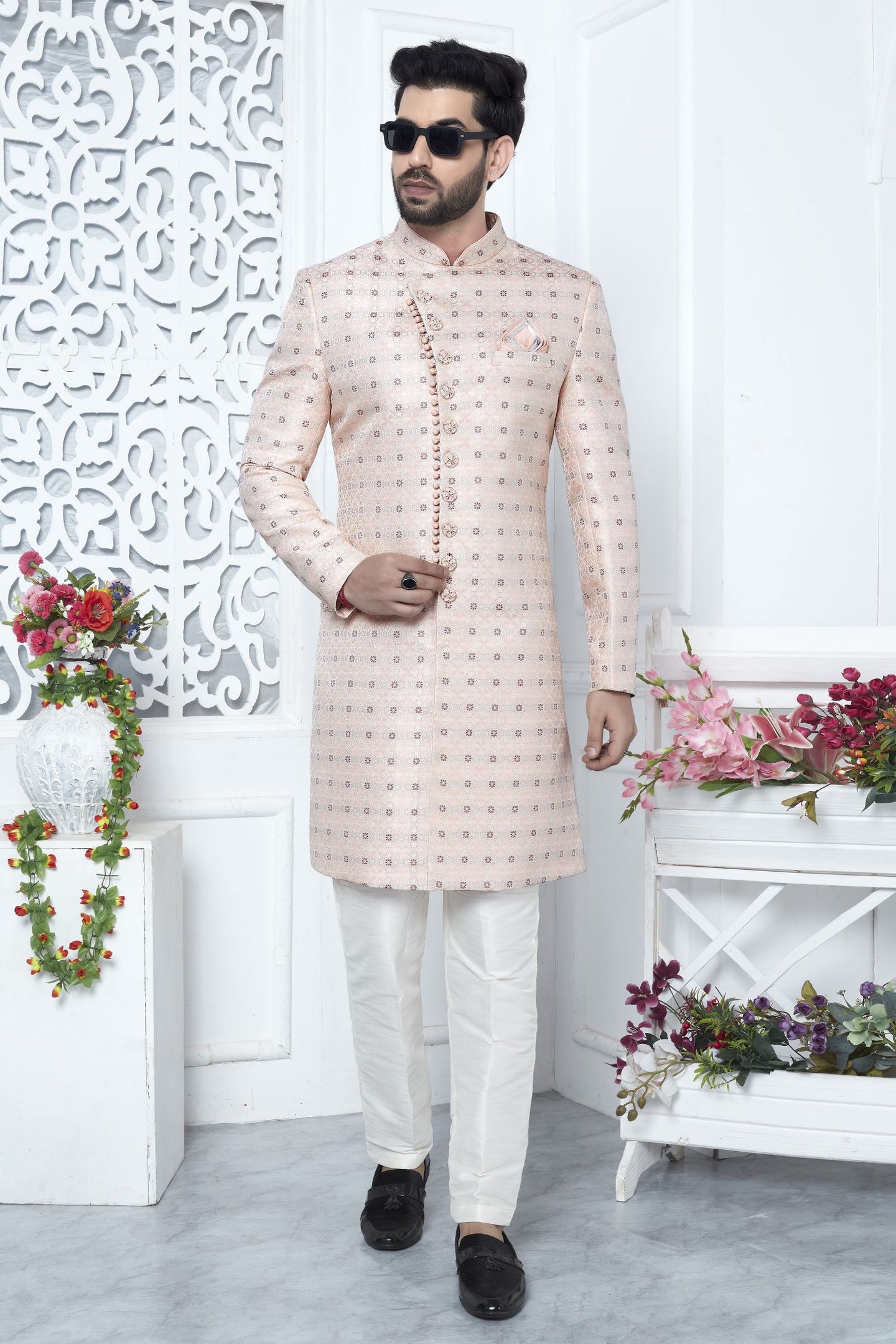 A man modeling a light pink sherwani with small gold geometric patterns, paired with off-white trousers and black loafers, standing in a decorated white room with floral arrangements.