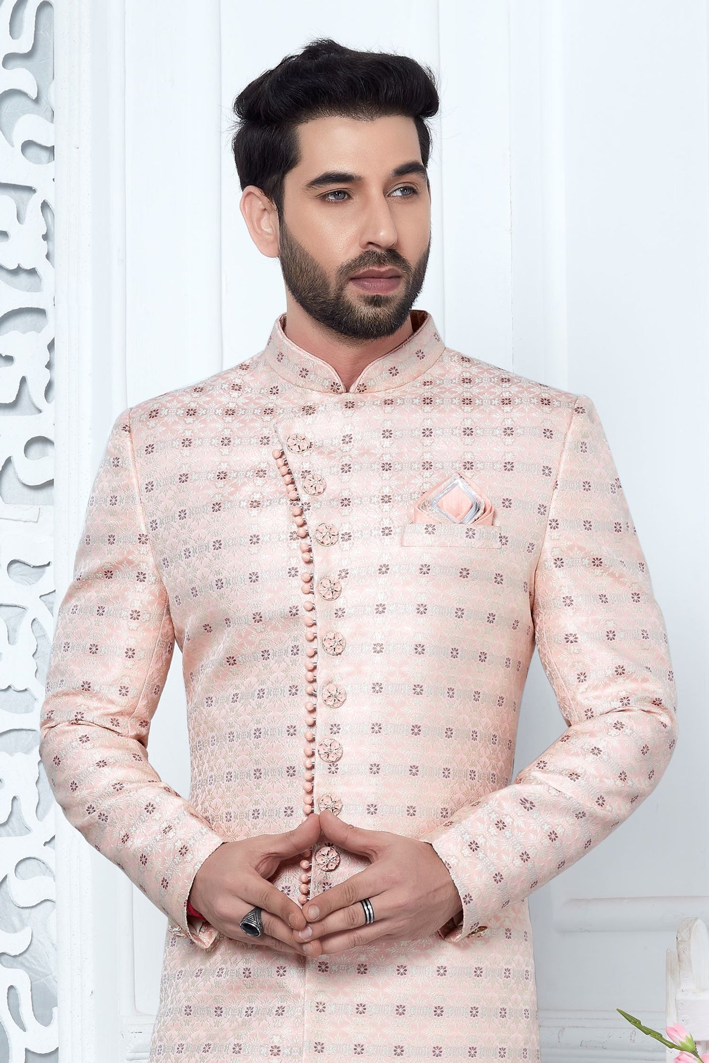 A man wearing a light peach-beige sherwani jacket with a small geometric pattern, standing against a white paneled wall.