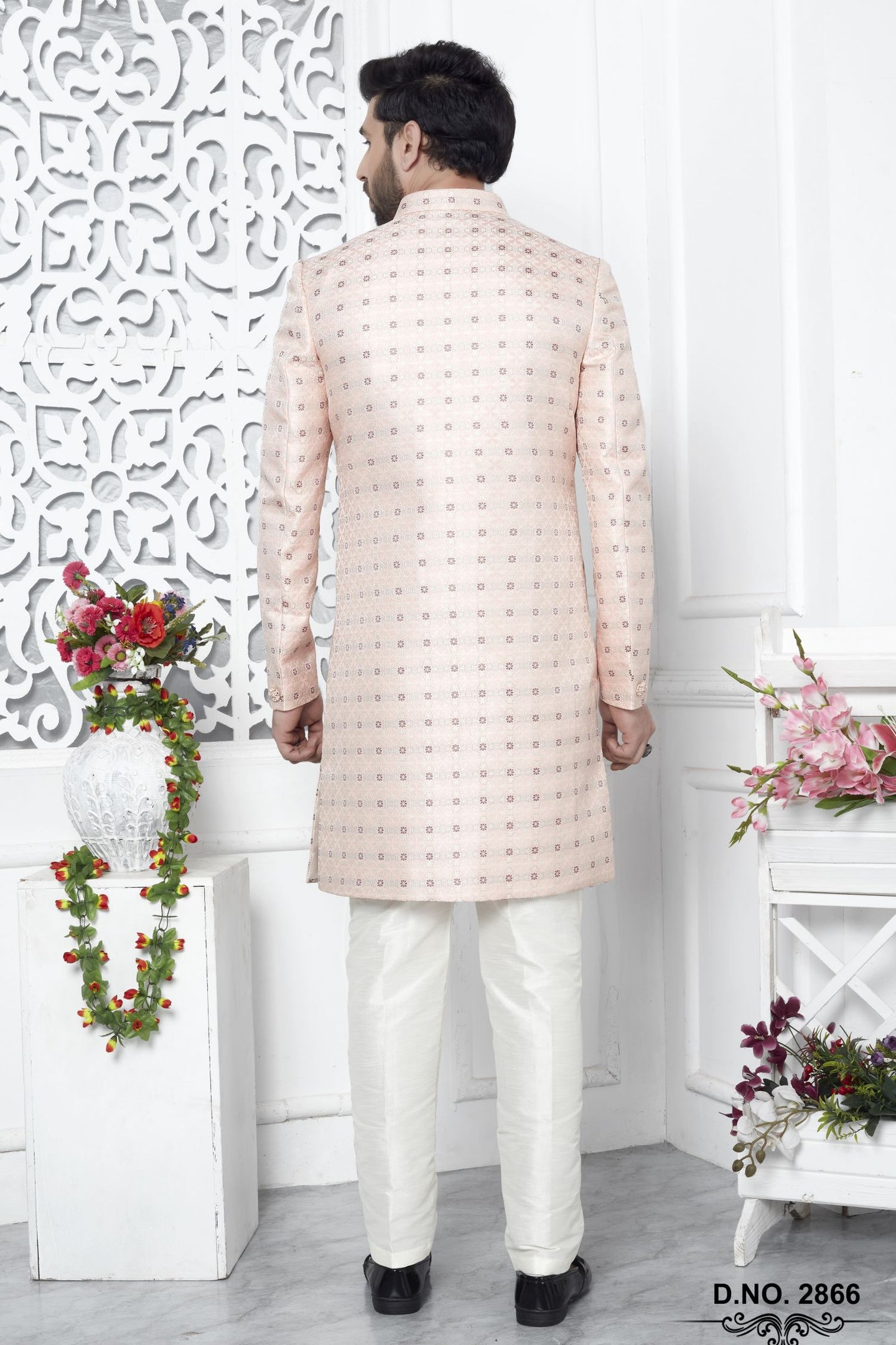 A man viewed from the back wearing a light peach-beige sherwani with a small repeating pattern and white trousers, standing in a decorated indoor setting with white walls and floral arrangements.