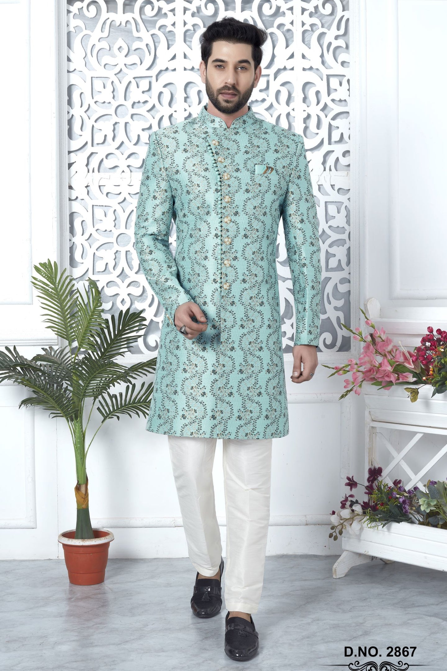 A man modeling a mint green embroidered sherwani with white trousers and black loafers, standing in front of a white decorative screen with potted plants on either side.