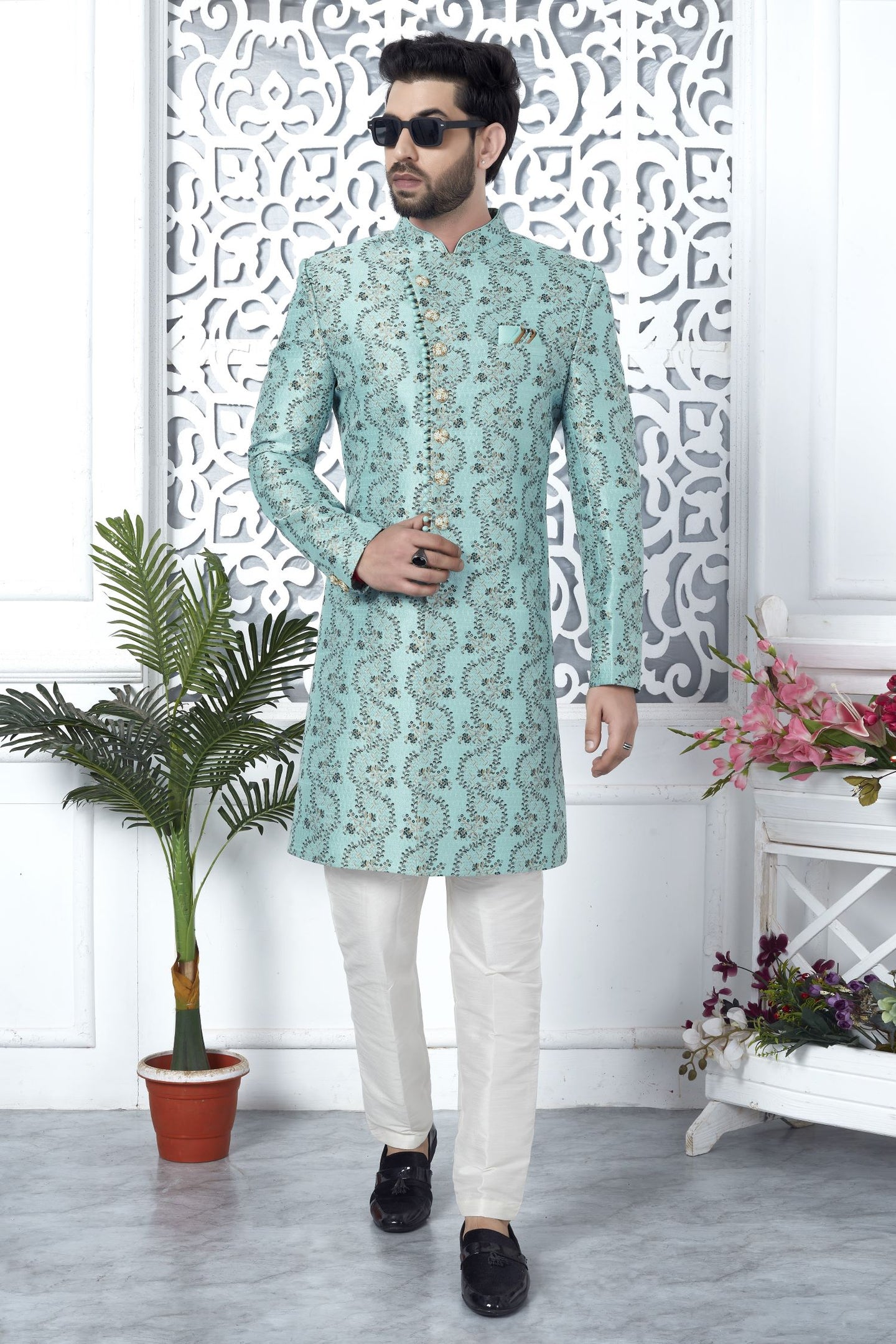 A man modeling a mint green embroidered kurta with white trousers and black loafers, standing in front of a white decorative screen with potted plants.