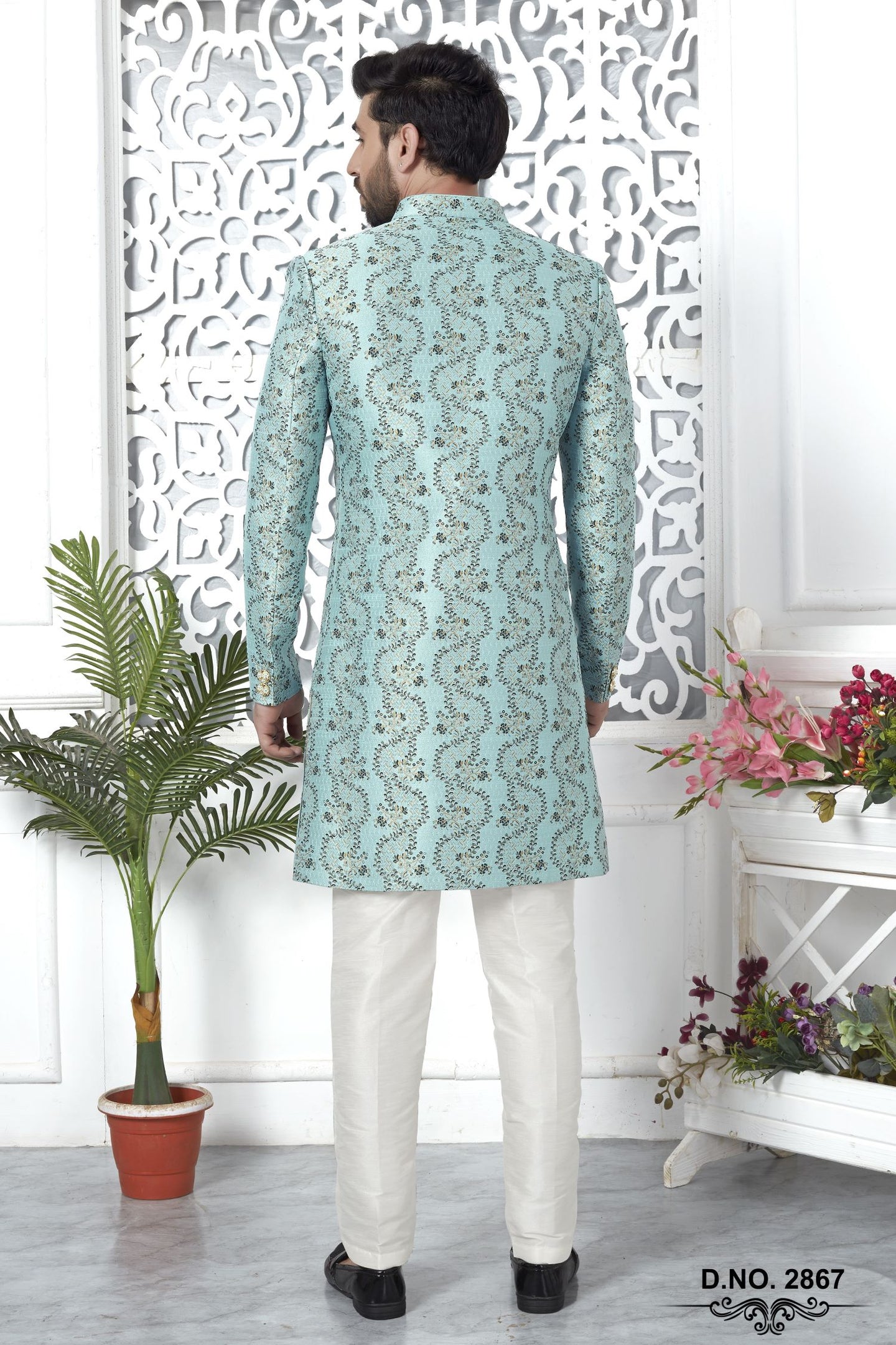 A man viewed from the back wearing a mint green embroidered sherwani with white trousers, standing in a decorated room with a white carved screen, a potted plant, and flowers.