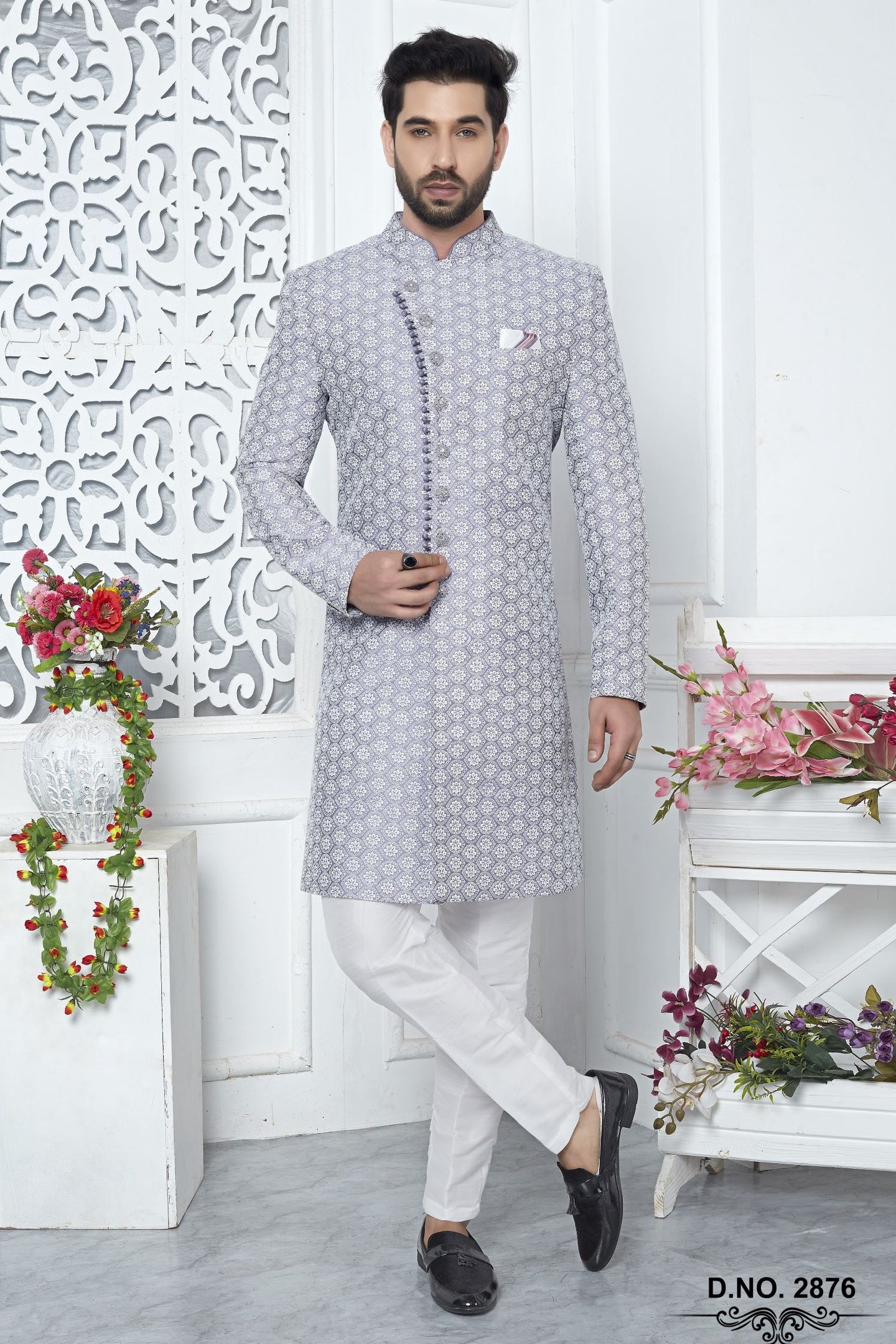 A man modeling a light gray patterned kurta with white trousers and black loafers, standing in a decorated indoor setting with white walls, floral arrangements, and a carved screen.