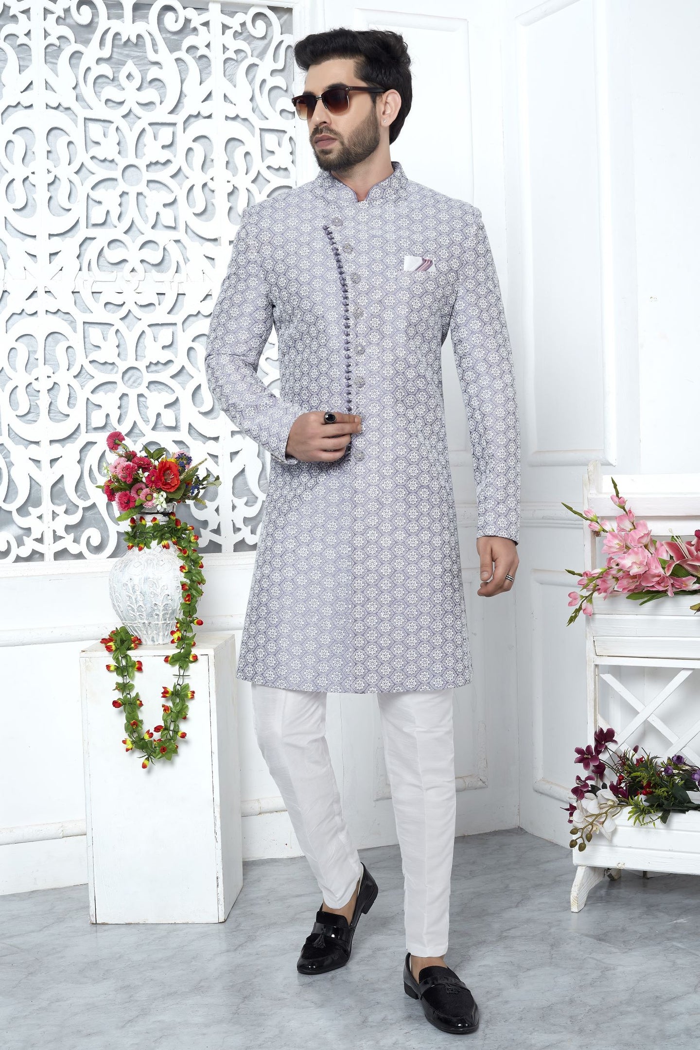 A man modeling a light gray kurta with a white geometric pattern, paired with white trousers and black loafers, standing in a decorated white room.