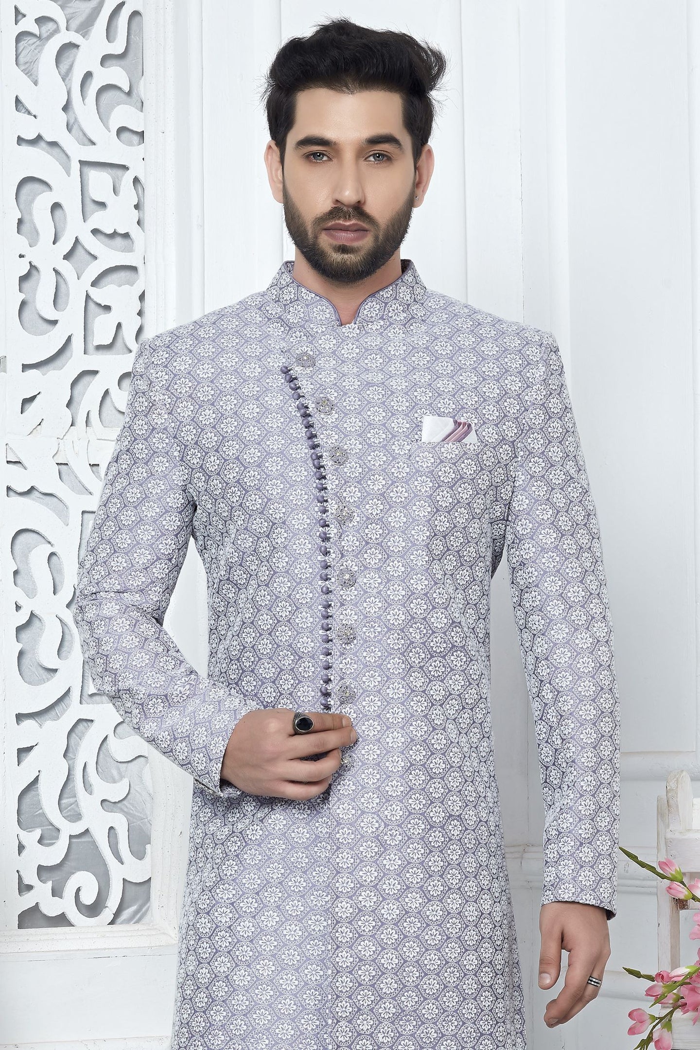 A man wearing a light gray and white patterned sherwani jacket with a mandarin collar, standing in front of a white decorative screen.