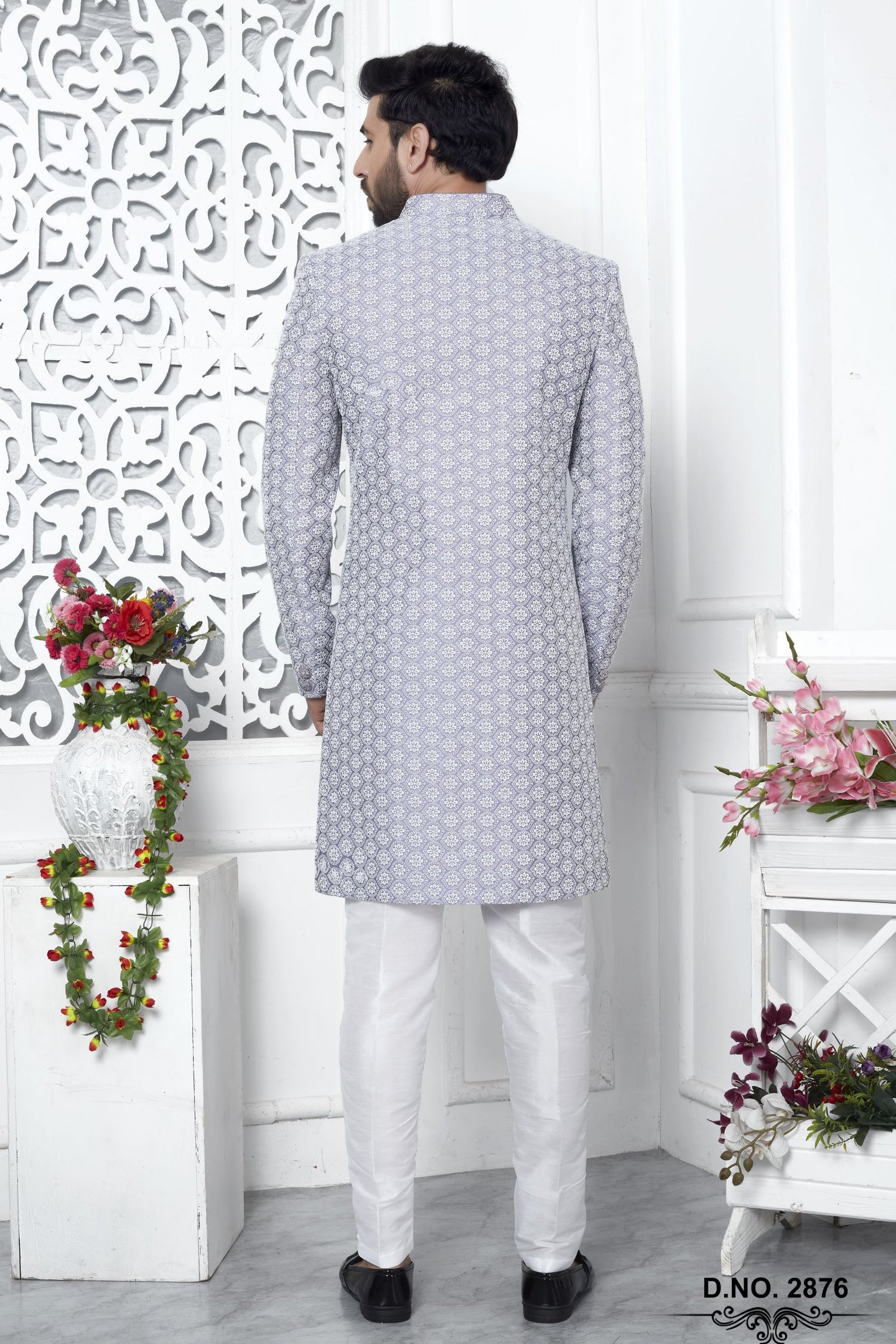 A man viewed from behind wearing a light gray patterned sherwani jacket with white trousers, standing in a decorated white interior with floral arrangements and a carved screen.