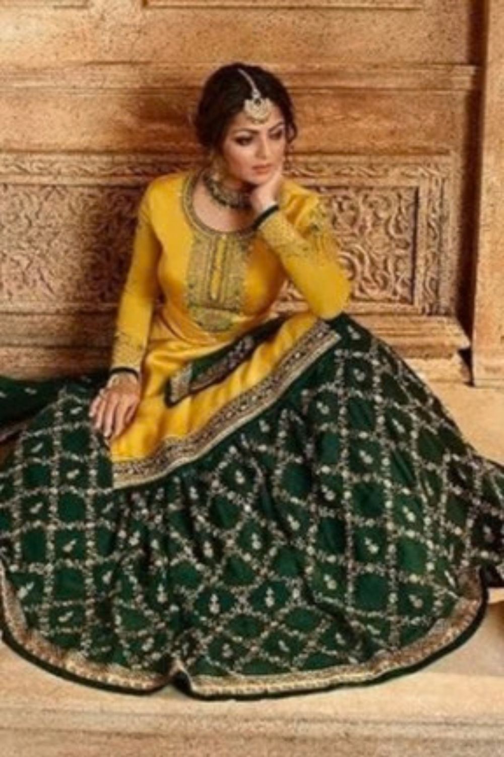 A full-length seated portrait shows a woman in a yellow embroidered haldi outfit with a dark green patterned skirt and traditional jewelry, reflecting haldi outfits for women, a haldi dress