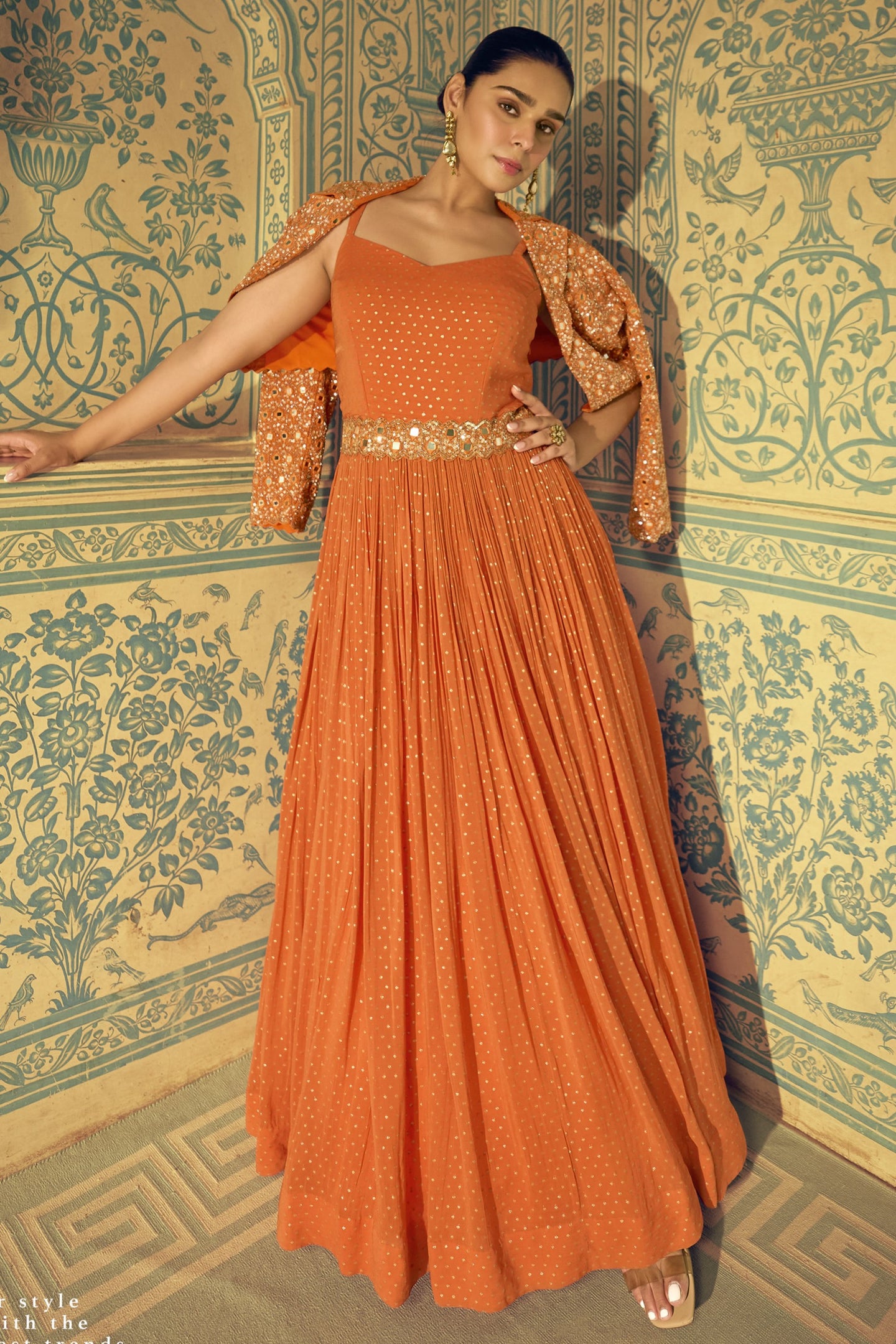 Woman wearing an orange dress with a gold belt against a decorative wall.