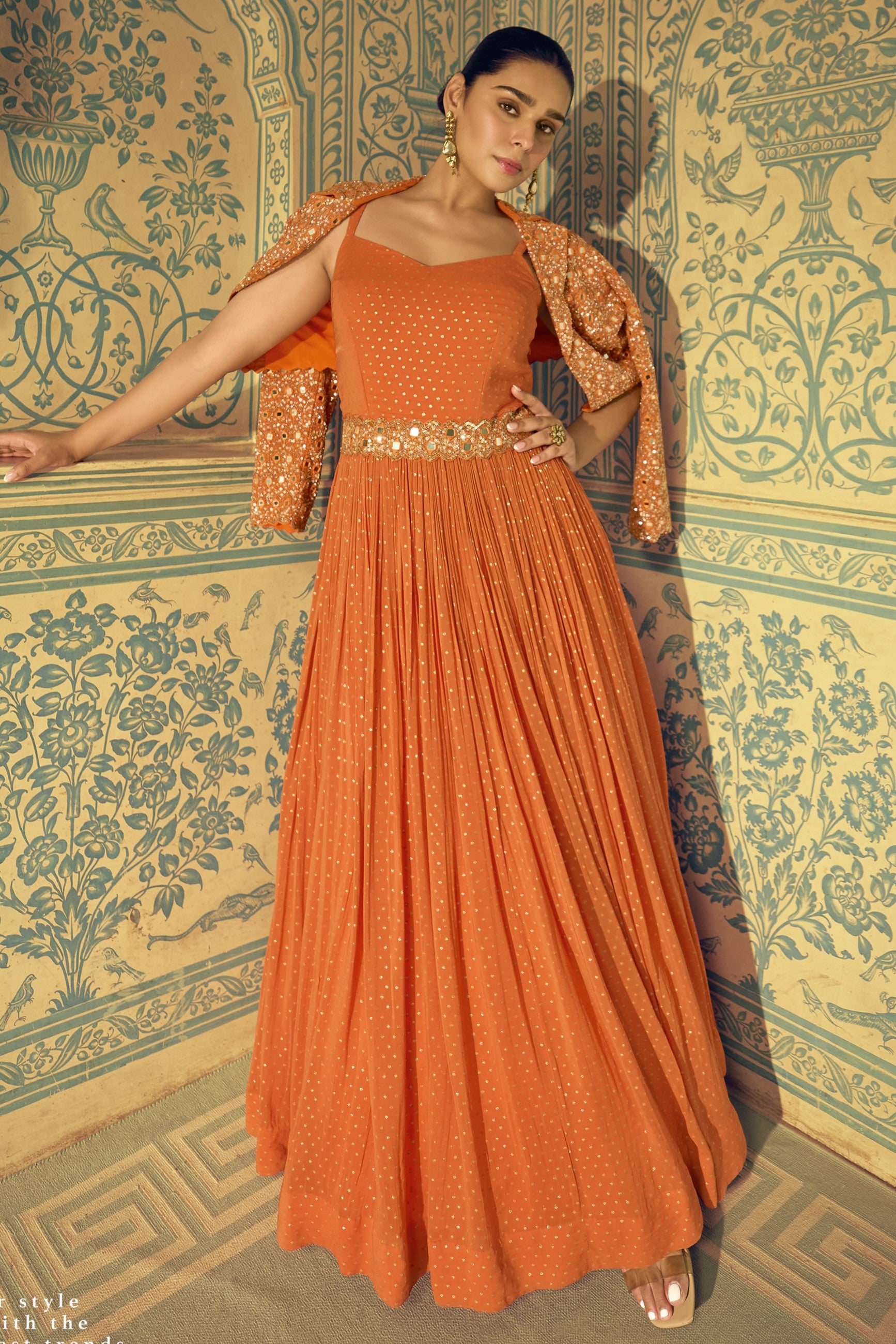 Woman wearing an orange dress with a gold belt against a decorative wall.