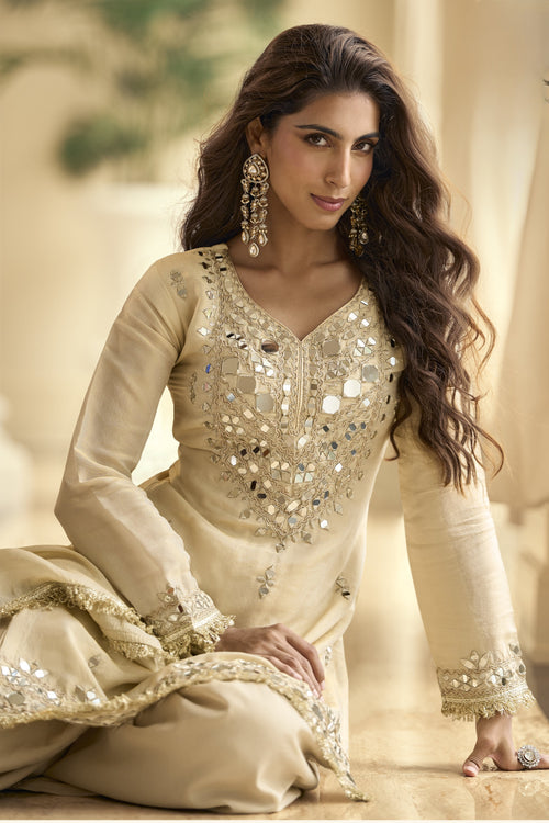 Woman sitting down wearing beige designer Plazo Suits for Party with real mirror work on the bodice and sleeves.