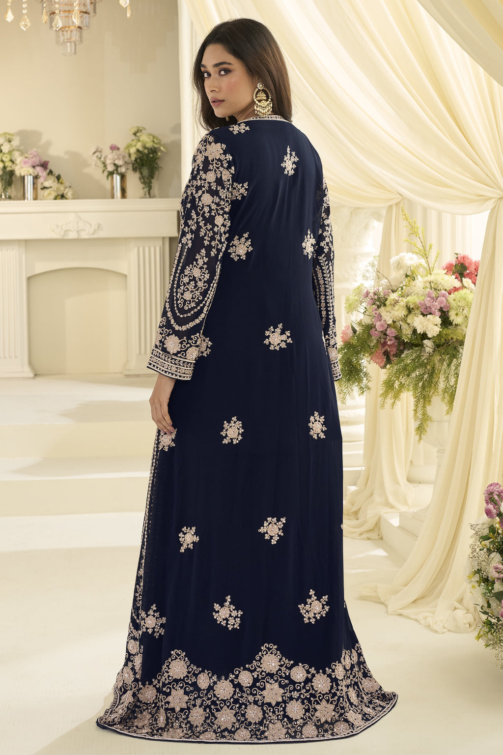 A rear-view portrait shows a woman in a navy blue embroidered ladies anarkali suit posing beside cream drapes and floral arrangements.