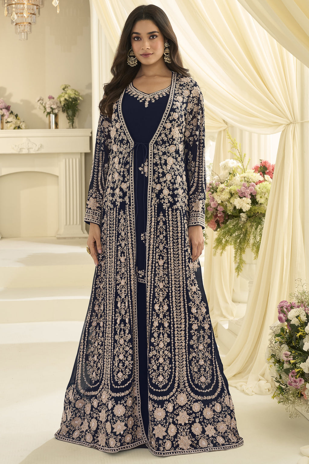 A woman poses for a full-length portrait in a navy blue Anarkali with jacket featuring ornate embroidery, surrounded by cream drapes and floral arrangements.