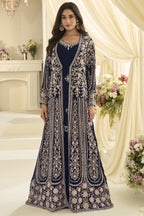 A woman poses for a full-length portrait in a navy blue Anarkali with jacket featuring ornate embroidery, surrounded by cream drapes and floral arrangements.