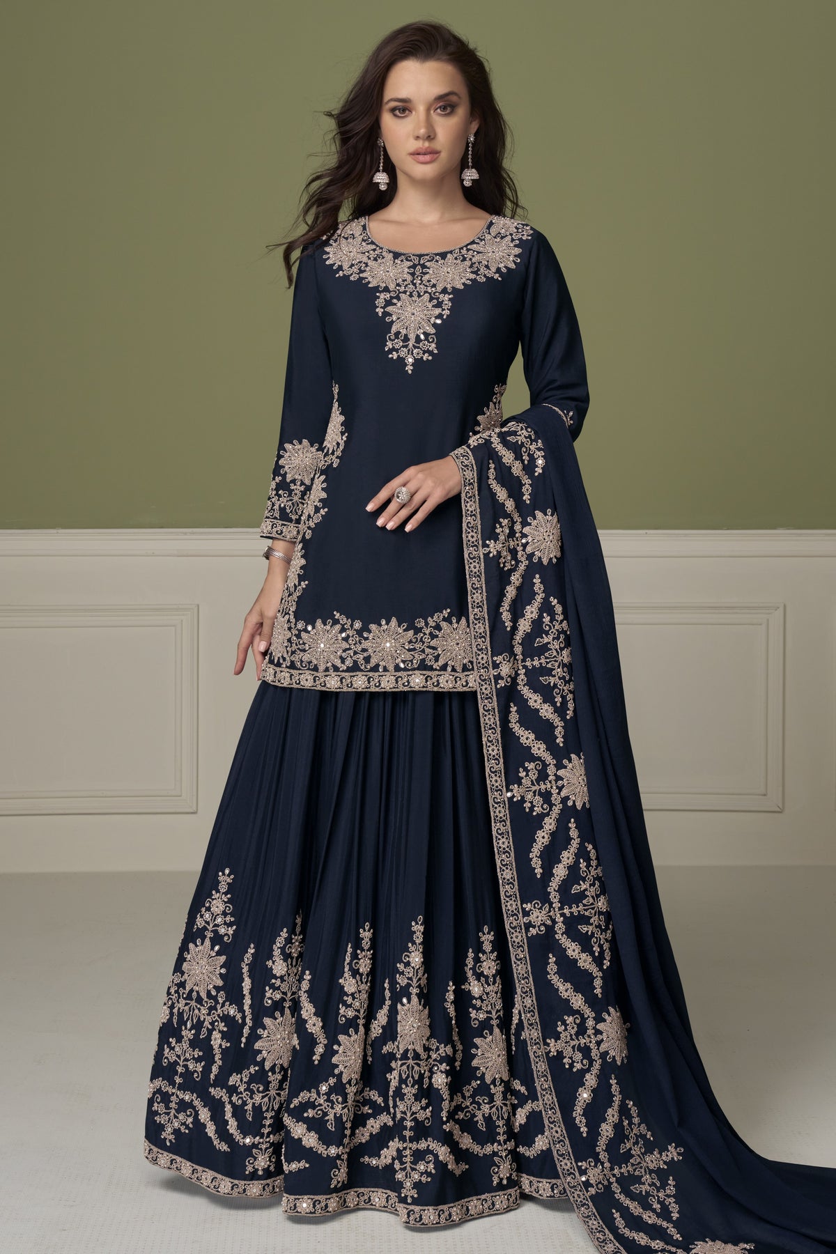 A woman modeling a navy blue traditional Indian outfit with extensive embroidery, featuring a long kurta, flared skirt, and matching dupatta against an olive green and white paneled studio background.