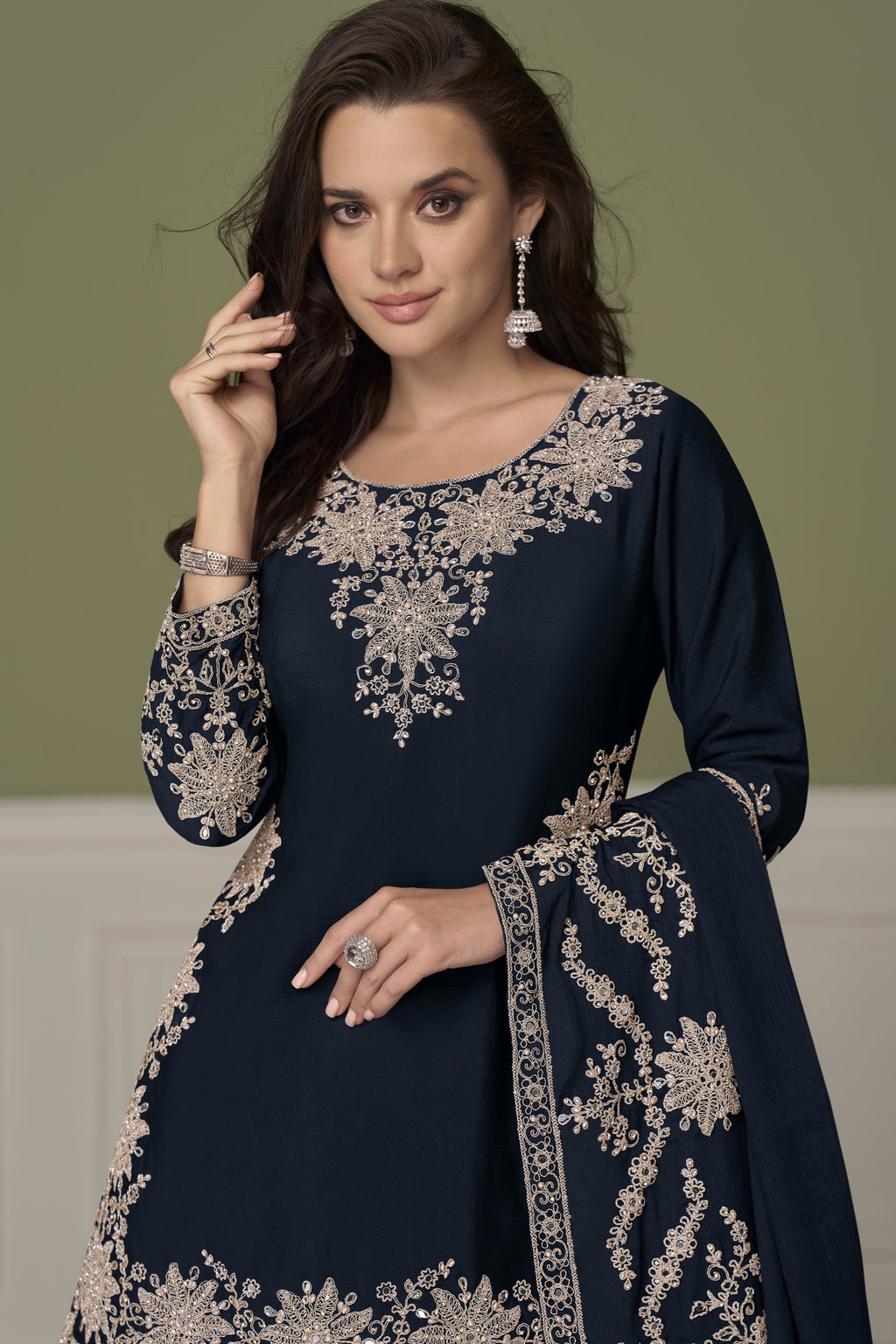 A woman modeling a navy blue traditional outfit with intricate embroidery, paired with a matching dupatta, set against a muted olive green background.