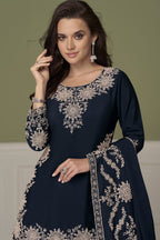 A woman modeling a navy blue traditional outfit with intricate embroidery, paired with a matching dupatta, set against a muted olive green background.