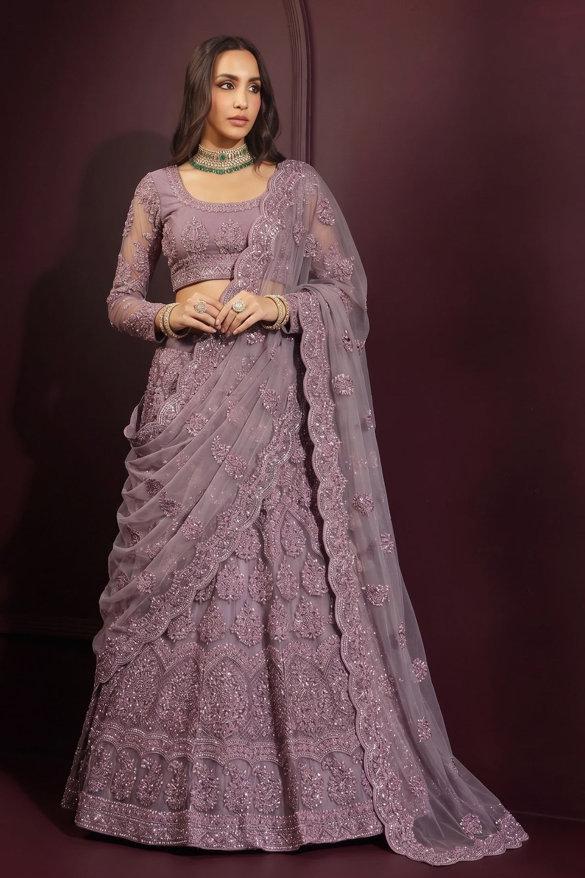 Model wearing a mauve lehenga choli for wedding with a long-sleeve embroidered blouse, flared net skirt and sheer dupatta draped over one arm.